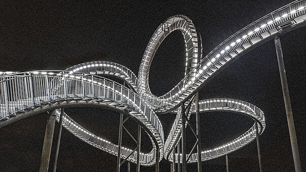 Detailansicht der hell erleuchteten Landmarke Tiger & Turtle bei Nacht. Die Lichter zeichnen die verschlungenen Wege der Achterbahn-Skulptur als leuchtendes Kunstwerk gegen den dunklen Himmel.