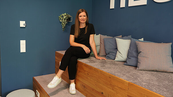 Lächelnd sitzt Denise Stahlhuth in schwarzem T-Shirt und Hose auf einer Holzbank mit grauer Polsterung, umgeben von blauen und grauen Kissen.