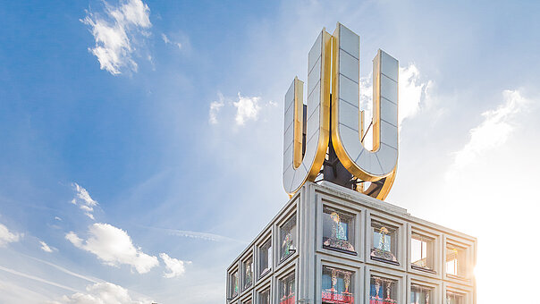 Froschperspektive auf das Dortmunder U. Das goldene U auf dem Dach des ehemaligen Brauereigebäudes strahlt vor einem blauen Himmel mit weißen Wolken.