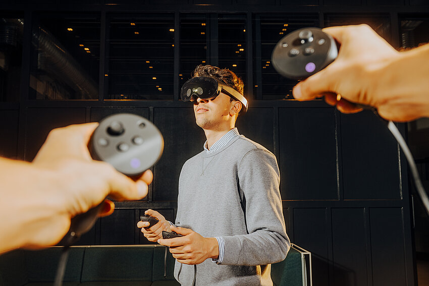 Aus der Ich-Perspektive werden zwei Controller auf einen Mann mit VR-Brille gerichtet. Er steht in einem dunklen Raum und hält ebenfalls Controller in den Händen.