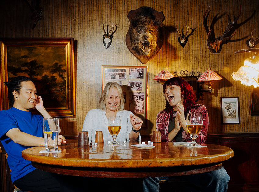 Ein Mann und zwei Frauen sitzen lachend an einem runden Holztisch in einer rustikalen Kneipe. An der Wand hängen Jagdtrophäen und Bilder.