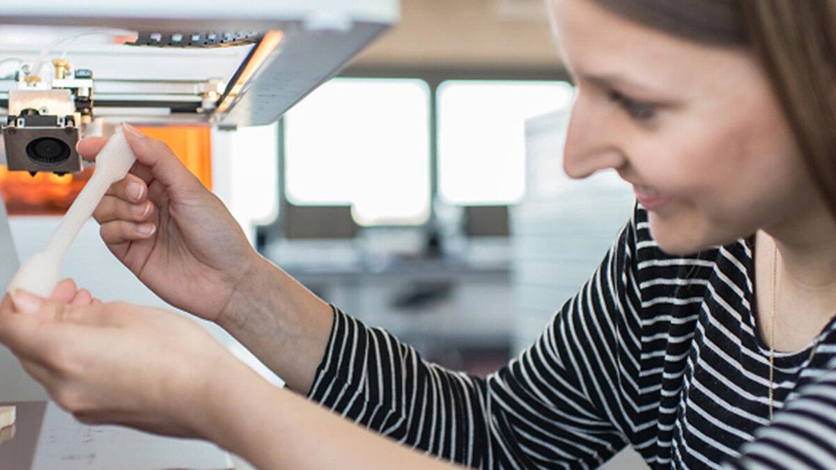 Eine junge Frau im gestreiften Hemd begutachtet lächelnd ein weißes, frisch gedrucktes 3D-Objekt. Sie entnimmt das Werkstück aus dem 3D-Drucker.