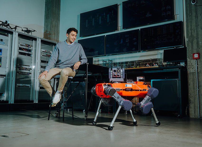 Ein Mann sitzt in einem Raum mit Computern. Vor ihm steht ein Roboterhund.