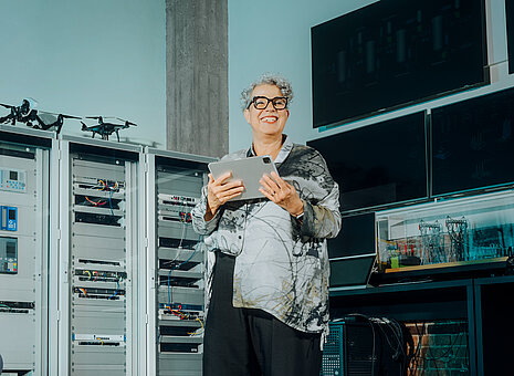 Eine Frau lächelt und hält ein Tablet in ihren Händen. Im Hintergrund hängen Computer-Bildschirme an der Wand.