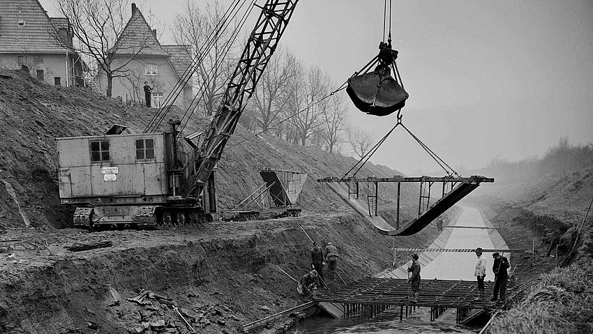 Ein Schwarz-Weiß-Bild zeigt Bauarbeiten an der Emscher. Ein großer Bagger hebt ein Bauteil an, während Arbeiter in der Grube stehen und an der Kanalisierung des Flusses arbeiten.