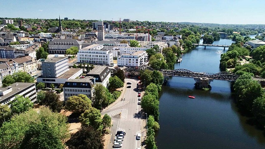 Luftaufnahme der Stadt Mülheim, die sich am Ufer der Ruhr erstreckt. Die Ansicht von oben zeigt die dichte Bebauung, grüne Alleen und die Weite des Flusses an einem sonnigen Tag.