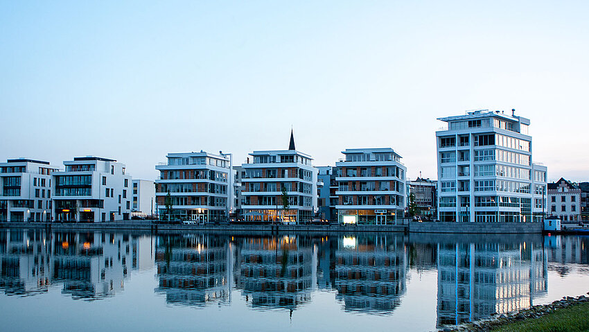 Eine Reihe moderner, weißer Wohngebäude säumt das Ufer. Die beleuchteten Fenster und die Architektur der Häuser spiegeln sich klar im ruhigen Wasser des Phoenix Sees, was eine stimmungsvolle, urbane Atmosphäre erzeugt.