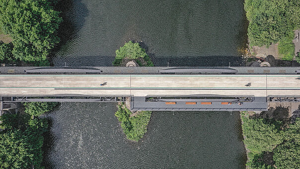 Luftaufnahme der Brücke des Radschnellwegs Ruhr, die einen dunklen Fluss überquert. Einzelne Personen sind auf dem hellen Weg zu sehen, der von grünen, baumbewachsenen Ufern gesäumt wird.