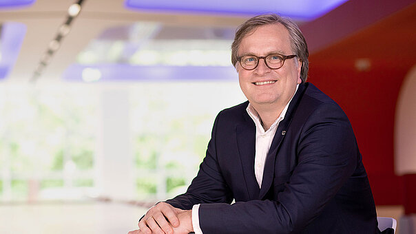 Ein Mann mittleren Alters (G DATA-Mitgründer Andreas Lüning) mit Brille und dunklem Sakko sitzt an einem Tisch in einem modernen, hellen Büro. Er hat die Hände vor sich gefaltet und blickt lächelnd in die Kamera.