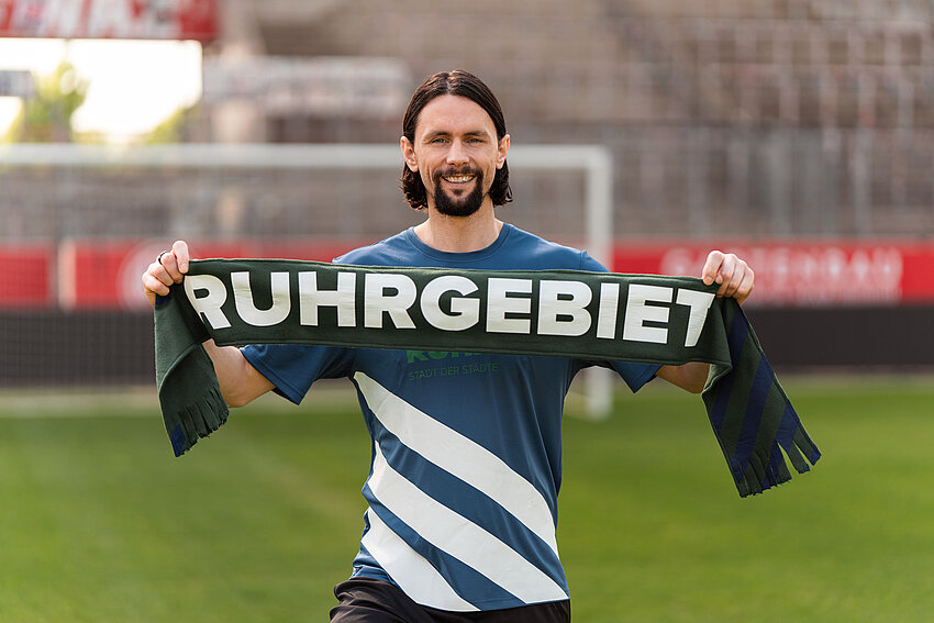 Der lächelnde Fußballprofi Neven Subotić steht auf einem Fußballplatz und hält einen Fanschal mit der weißen Aufschrift „Ruhrgebiet“ in die Kamera.