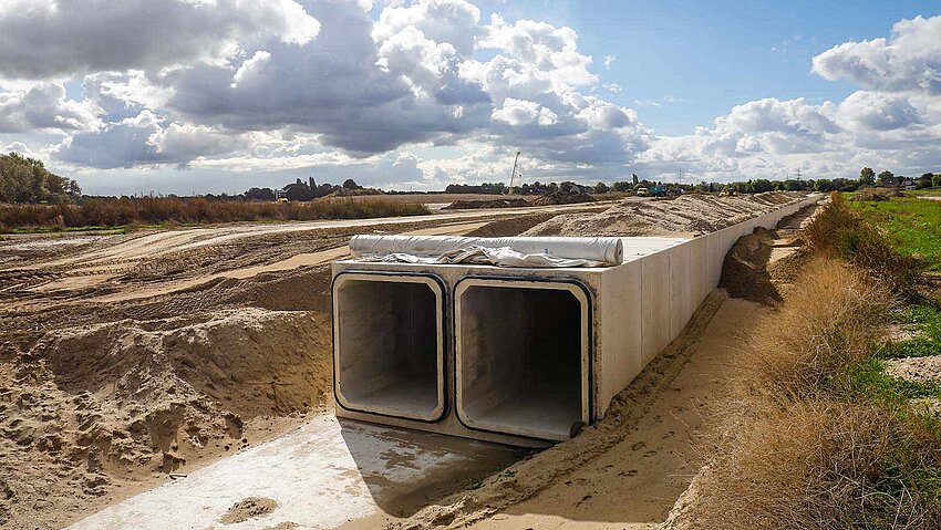 Eine Baustelle mit einem großen, rechteckigen Betonrohr, das in den Boden eingelassen ist. Ringsherum sind weitläufige Erdarbeiten zu sehen, mit aufgeschütteten Erdhügeln und Fahrspuren von Baufahrzeugen. Im Hintergrund sind weitere Baufahrzeuge und eine ländliche Landschaft unter einem bewölkten Himmel erkennbar.