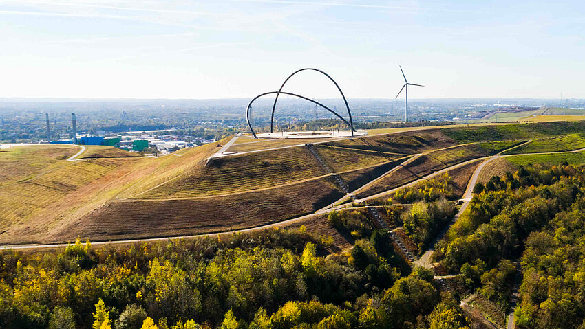 Weitläufiger Blick über eine begrünte Haldenlandschaft der Halde Hohewart. Auf dem Gipfel der Halde steht die große Stahlskulptur des Horizontobservatoriums, daneben ein Windrad. Am Horizont ist das Ruhrgebiet zu erkennen.