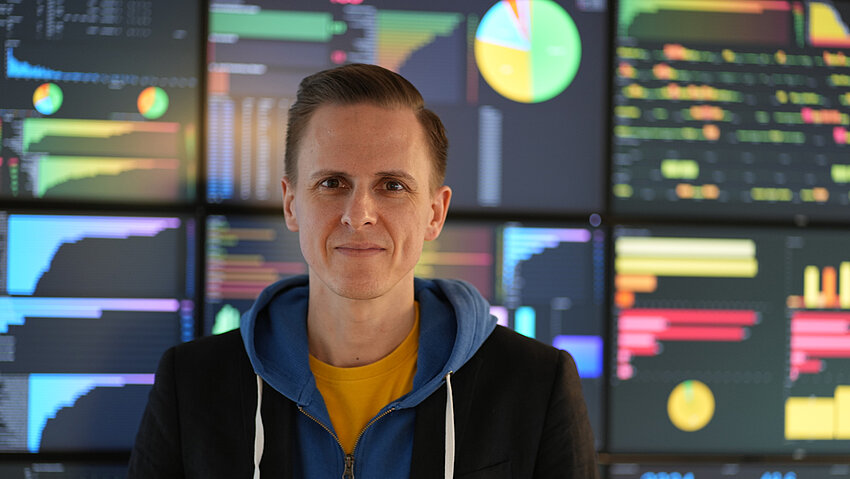 Porträt von Felix Nensa in einem Kontrollraum oder Datenzentrum. Der Mann trägt einen blauen Kapuzenpullover und ein Sakko. Hinter ihm zahlreiche Bildschirme mit bunten Graphen und Diagrammen.