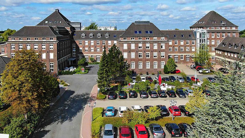 Das weitläufige Backsteingebäude mit Schieferdächern von G DATA wird von grünen Bäumen umrahmt. Auf dem großen Parkplatz im Vordergrund stehen zahlreiche bunte Autos unter blauem Himmel.