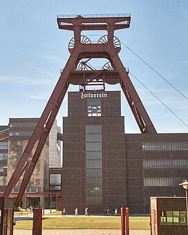Das rote Fördergerüst von Schacht 12, Wahrzeichen der Zeche Zollverein. Das imposante Industriedenkmal der Industriekultur im Ruhrgebiet steht vor einem dunklen Backsteingebäude.
