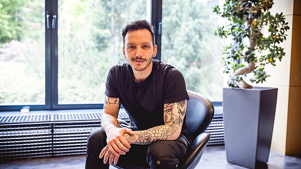 Alper Bate sitzt entspannt mit dunklem T-Shirt und tätowierten Unterarmen in einem schwarzen Sessel vor einem großen Fenster, das Bäume und Tageslicht zeigt.