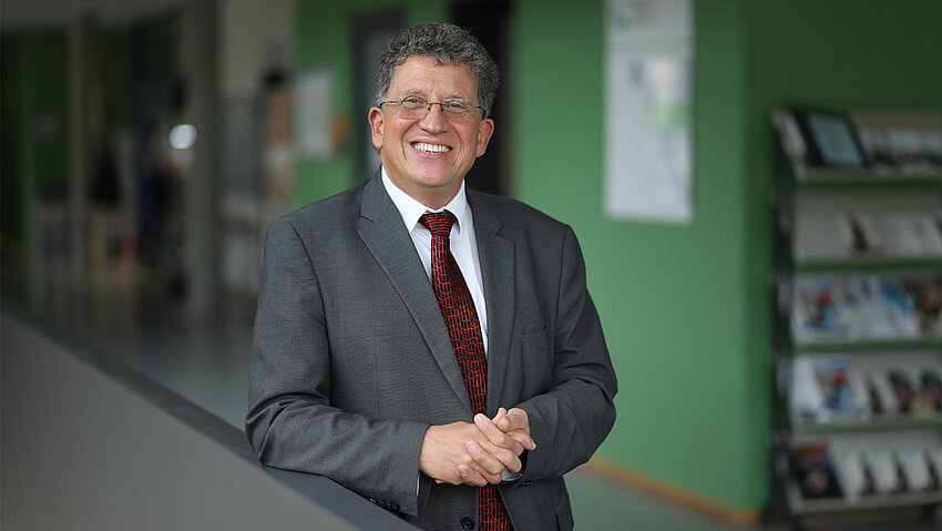 Michael Brodmann — ein Mann mittleren Alters mit Brille, grauem Anzug und rot gemusterter Krawatte — lehnt sich lächelnd an ein Geländer. Er hat die Hände gefaltet und blickt in die Kamera.