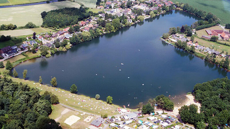 Eine weite Luftaufnahme des Ternscher Sees. An seinen Ufern liegen ein Campingplatz und eine kleine Siedlung, eingebettet in eine grüne Wald- und Wiesenlandschaft.