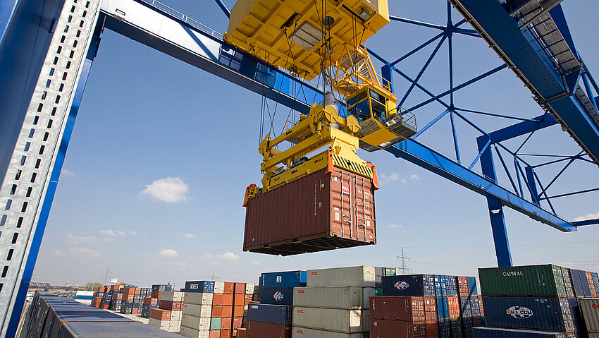 Eine große Containerbrücke verlädt bei sonnigem Wetter einen Schiffscontainer. Im Hintergrund sind zahlreiche weitere Container im Duisburger Hafen gestapelt.