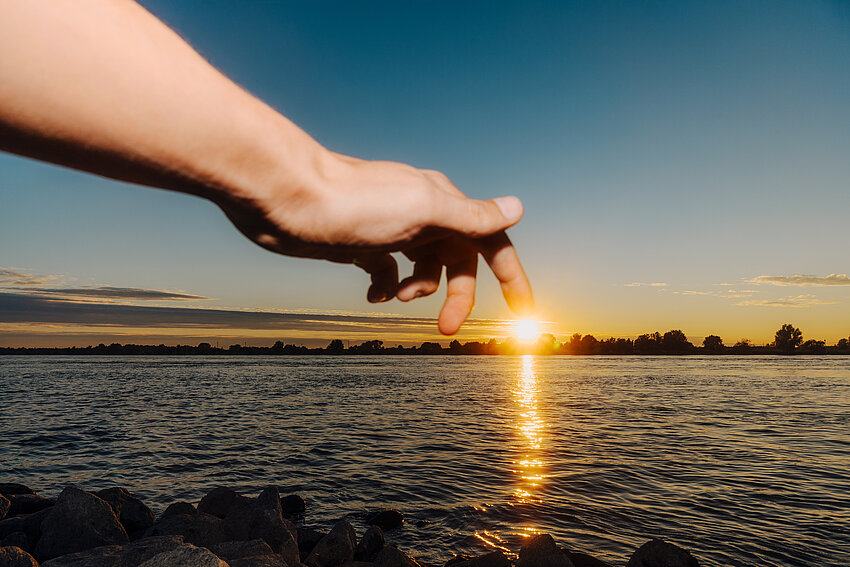 Zwei Finger einer Hand laufen symbolisch auf der Spiegelung der untergehenden Sonne auf dem Wasser. Am Horizont ist eine Stadtsilhouette unter klarem Himmel erkennbar.