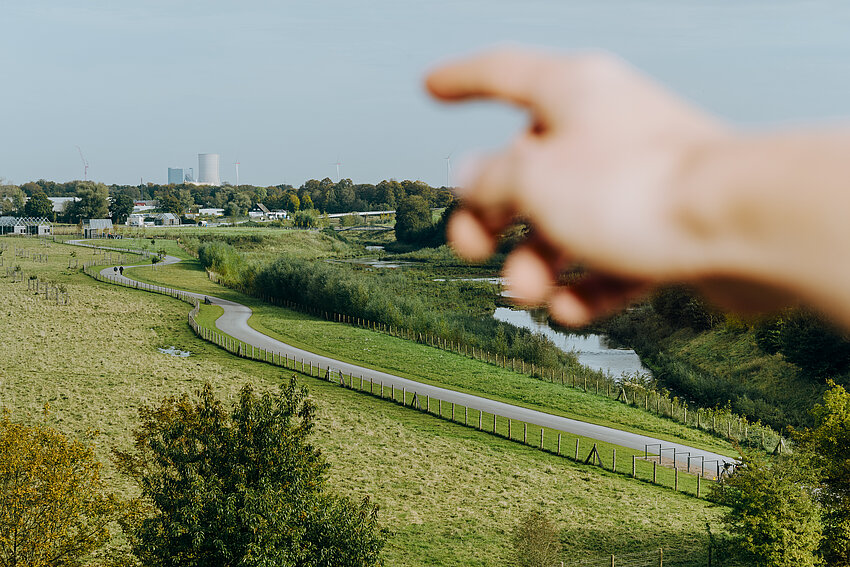 Aus der Ich-Perspektive zeigt eine Hand auf eine weite, grüne Landschaft mit einer Straße und einem Fluss. Am Horizont ist eine Industrieanlage zu erkennen.